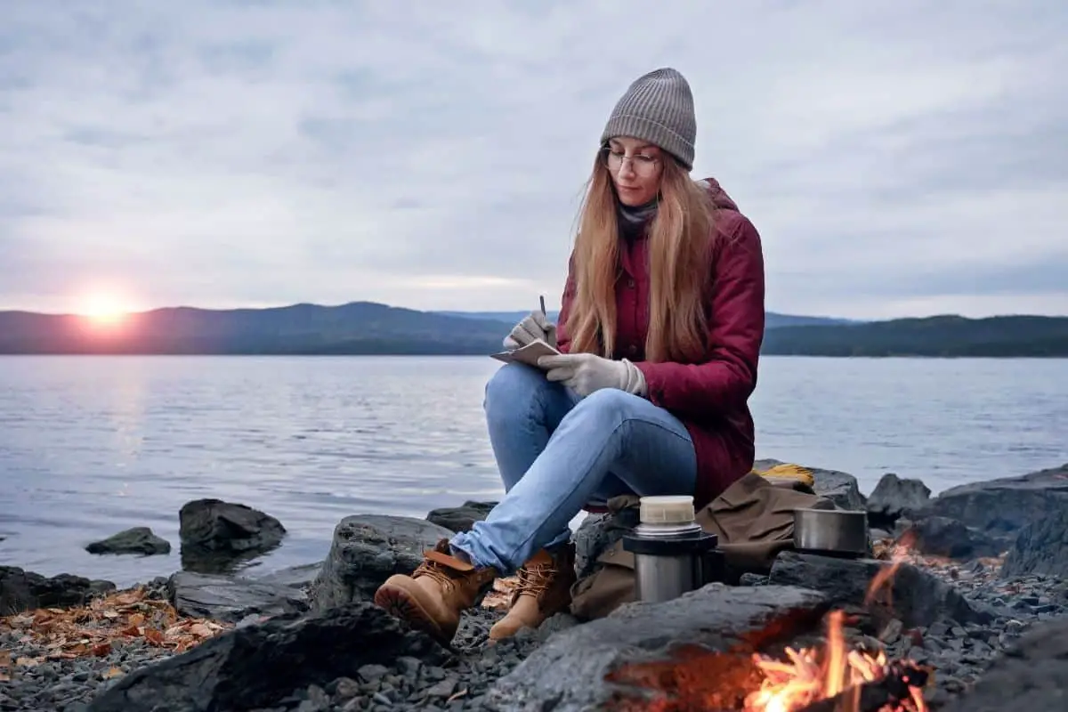 a woman sitting on rocks by the water writing in front of a fire