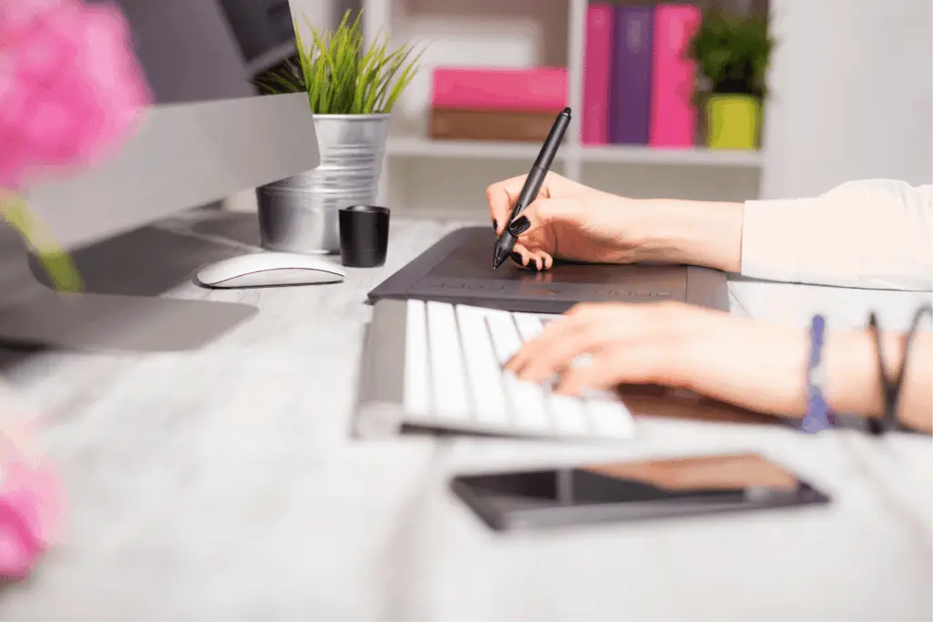 Woman with black painted fingernails typing on white keyboard with one hand and writing on electronic notebook with the other hand.