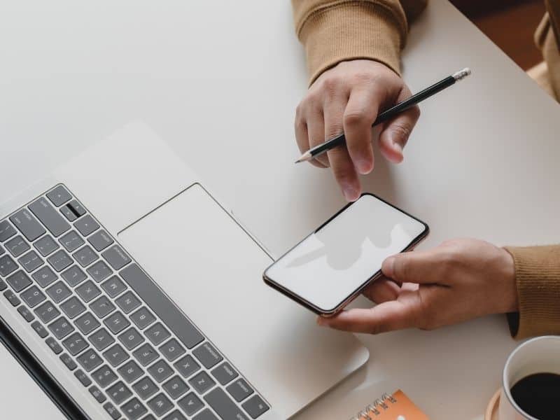 a person looking at their smartphone with a pen in hand in front of a laptop