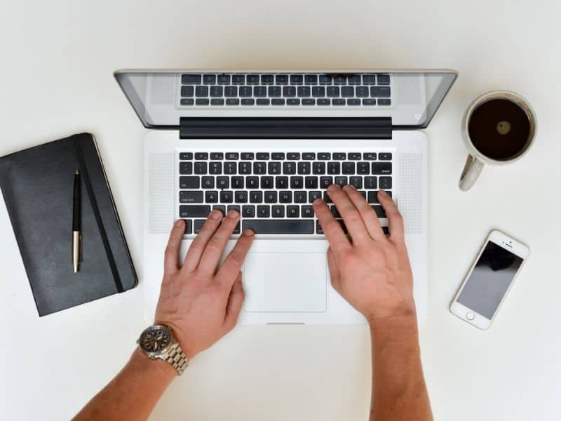 hand typing on a laptop computer with a notebook and cup of coffee on the table beside