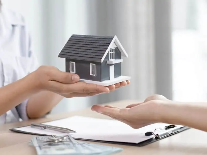 a pair of hands holding a model home over a desk with paperwork on it
