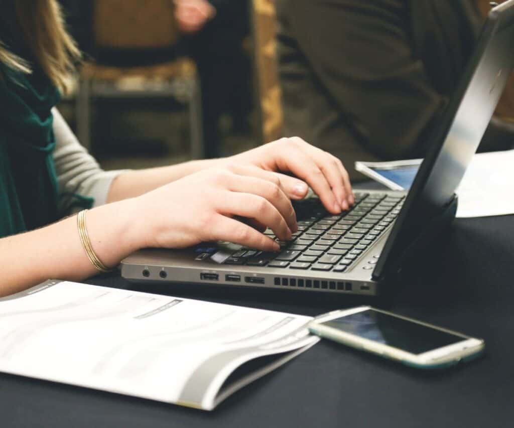 a woman typing on a laptop with a notebook and phone on the side