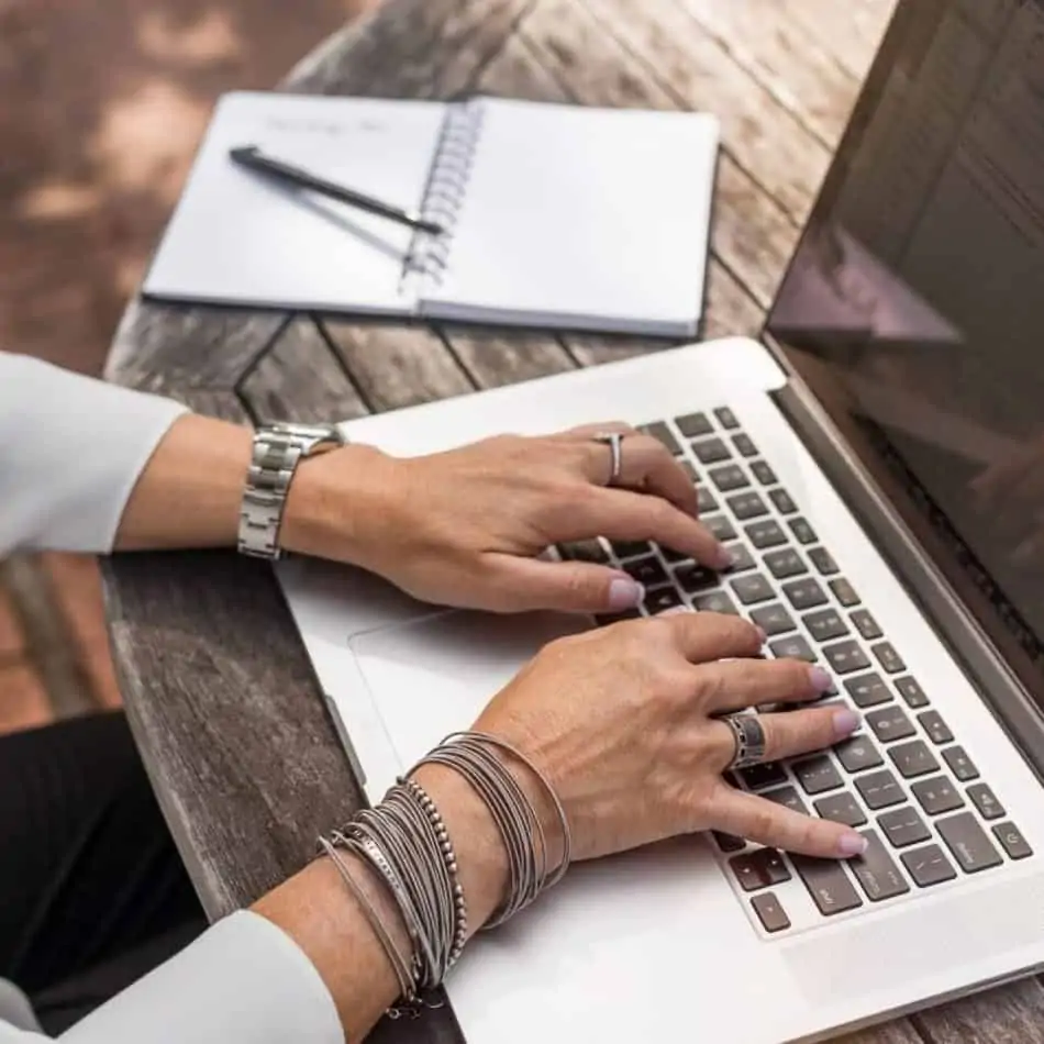 hands typing on a laptop with jewelry on and a notepad