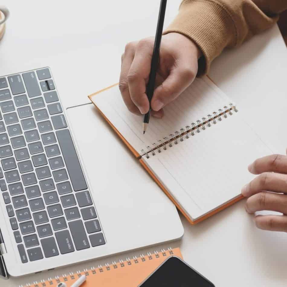 someone writing on a notepad in front of a laptop